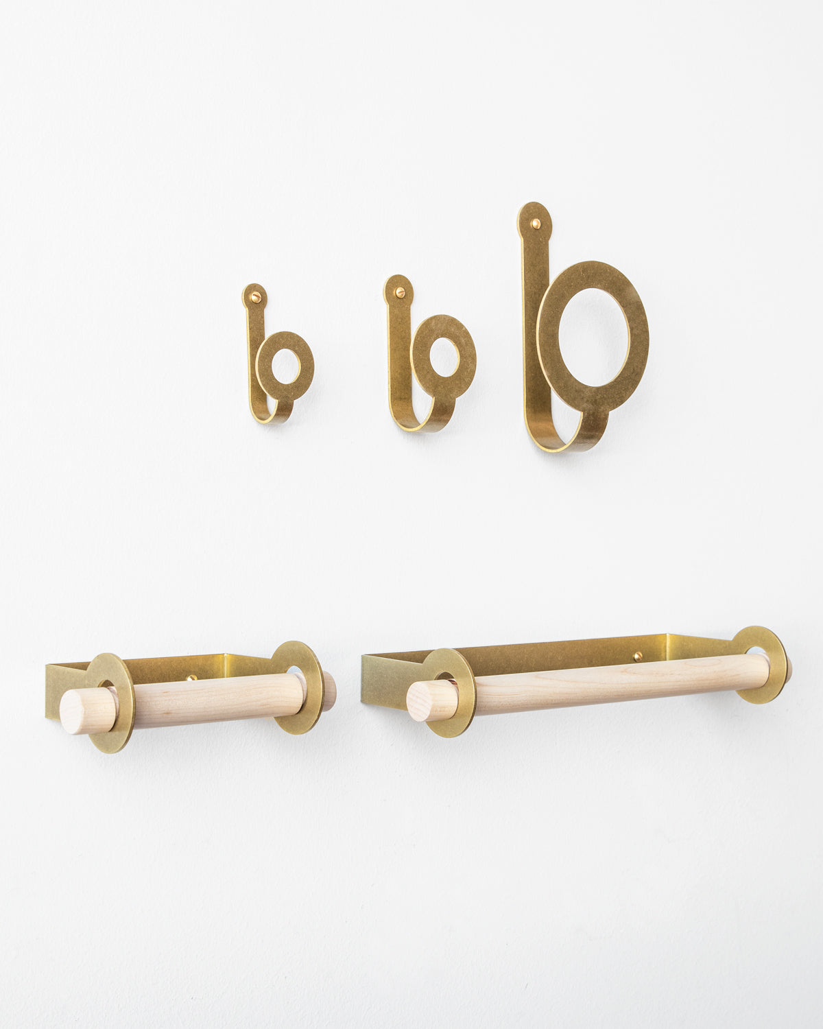 Three sizes of brass wall hooks above a brass toilet paper holder and brass paper towel holder, both empty. #finish_brass