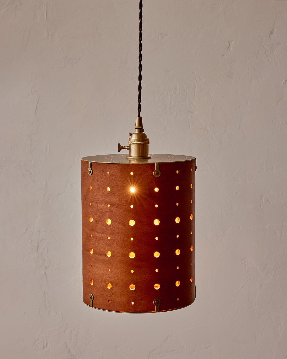 A cylindrical pendant light with a leather shade featuring perforations, suspended by a brass fitter and attached to a twisted cloth cord. #color_tan harness