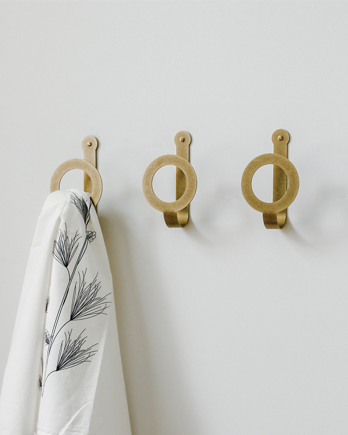 Three oversized brass wall hooks in a line. One holds a white towel patterned with black flowers. #finish_brass