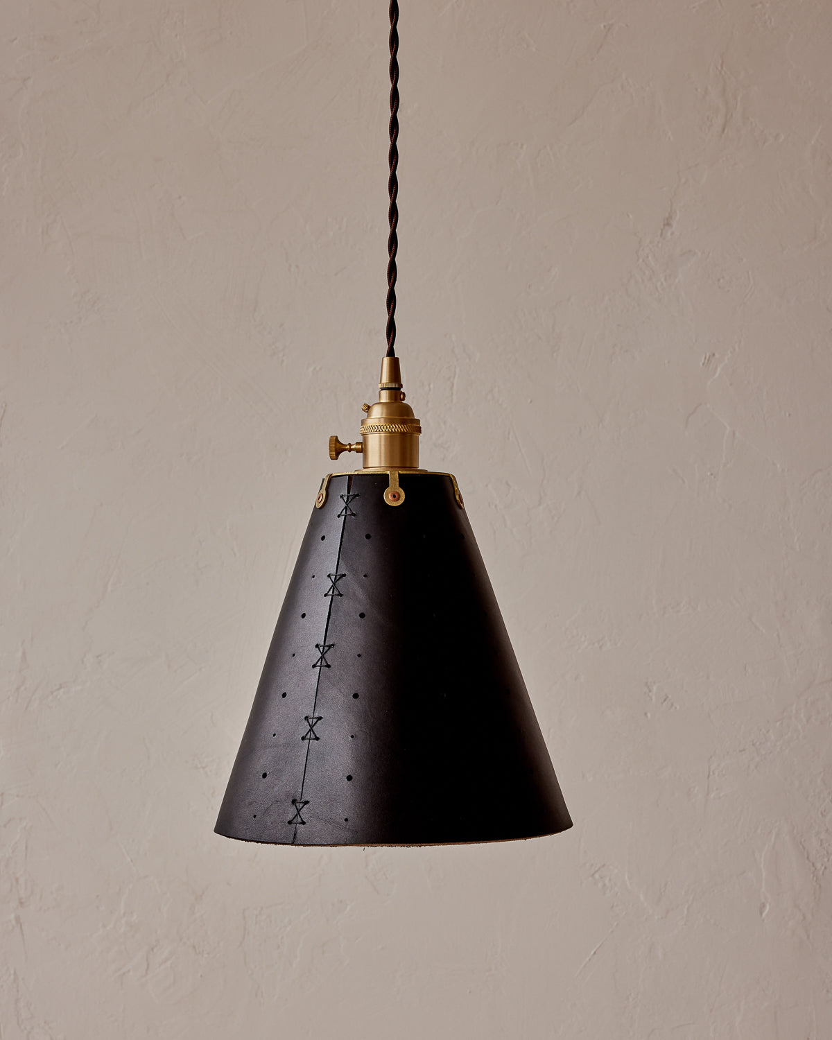 A conical pendant light with a black leather shade featuring patterned perforations, suspended by a brass fitter and a twisted cloth cord. #color_black harness
